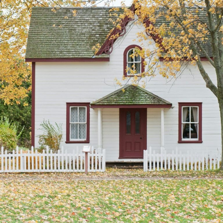 Weißes Haus mit rotem Dach und Türen, umgeben von herbstlichen, bunten Blättern.